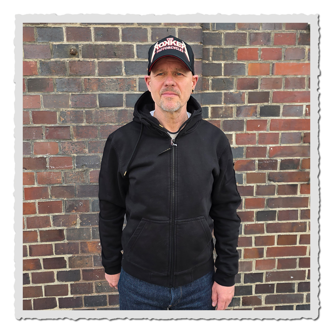 Person in schwarzem Zip Hoodie von vorn vor Backsteinwand, mit Kappe, dunkler Jeans und geradem Stand.